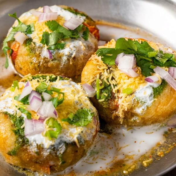 Three Indian chaat-filled puris topped with yogurt, chutneys, sev, red onion, cilantro, and green chilies on a metal plate.