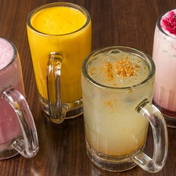 Four colorful frothy drinks in glass mugs sit on a dark wooden table, including pink, mango-yellow, pale green, and berry-topped beverages.